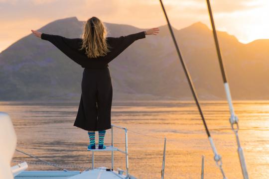 Lady in the front of the deck doing the Leonardo DiCaprio pose from Titanic with mountains and midnight sun in the background